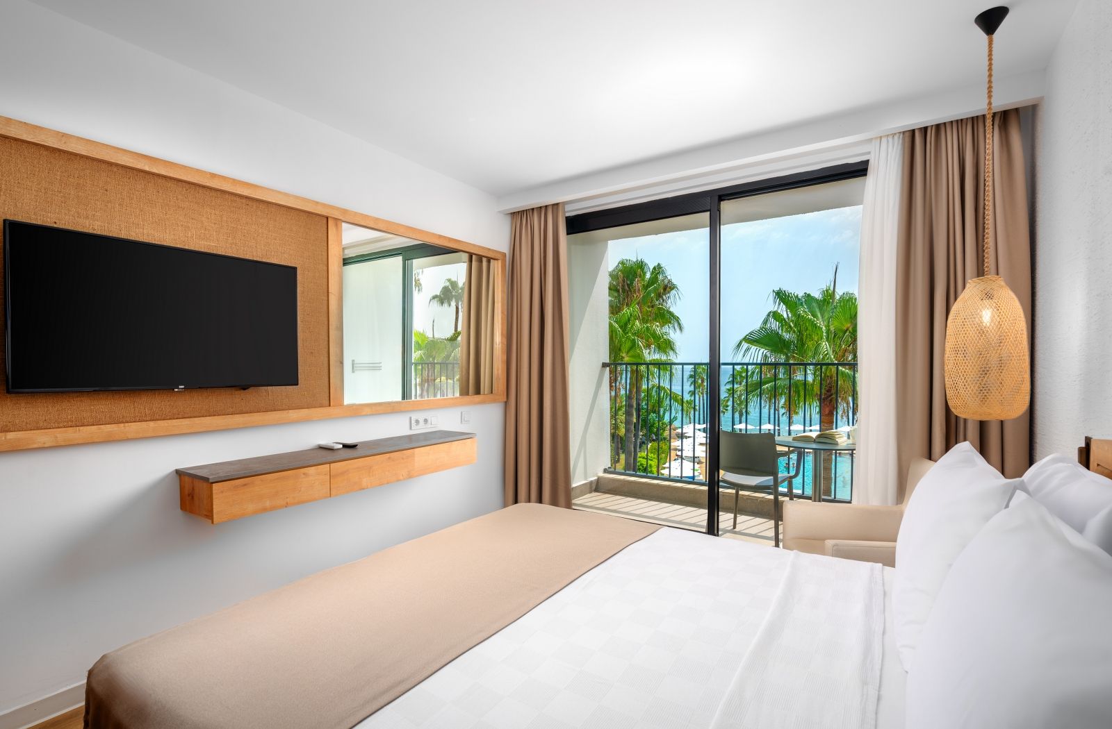 Camera d'albergo con un letto, una TV a muro, un tavolino, un balcone vista piscina e un lampadario.