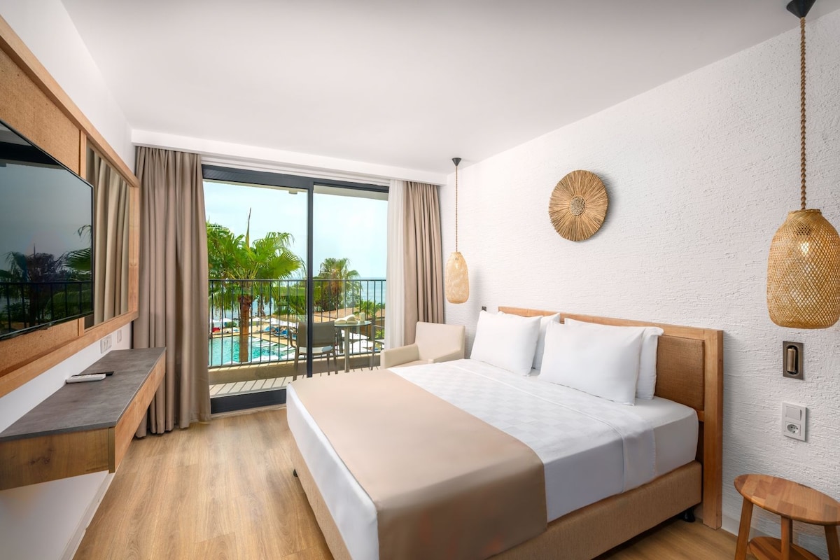 A hotel room with a bed, a TV, a desk, a balcony with a pool view, and a hanging light fixture.