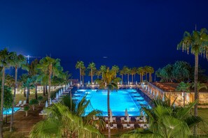 A nighttime view of a rectangular swimming pool surrounded by palm trees and lounge chairs, with a view of the ocean in the background. - Yalihan Aspendos Hotel (Alanya)