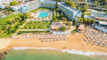 Vue aérienne d’un complexe hôtelier comprenant une piscine, une plage avec des transats et un ensemble de bâtiments.