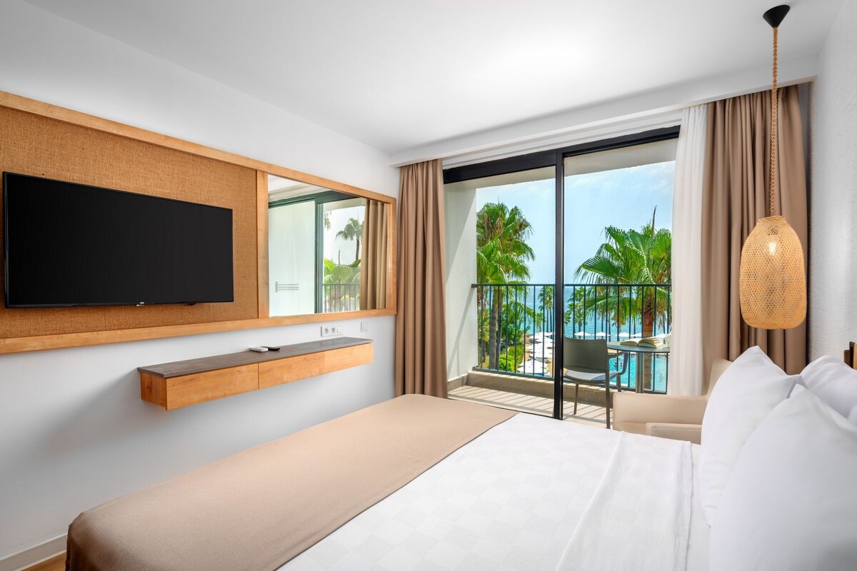 A hotel room with a bed, a mounted TV, a small table, a balcony with a pool view, and a hanging light fixture.