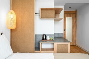 Une chambre d’hôtel moderne comprenant un lit, un petit bureau avec une bouilloire, une tasse et un plateau, un luminaire suspendu et une porte en bois.