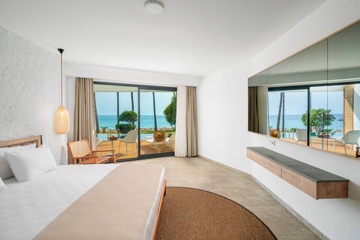 A modern hotel room with a large bed, a seating area with a view of the sea, and a large mirror reflecting the room.