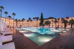 Zona de piscina de hotel con múltiples sillas de descanso y sombrillas, rodeada de palmeras y un edificio con balcones iluminados.