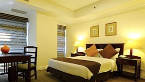 Una habitación de hotel con cama, mesitas de noche, una silla, una mesa y un ventanal con persianas.