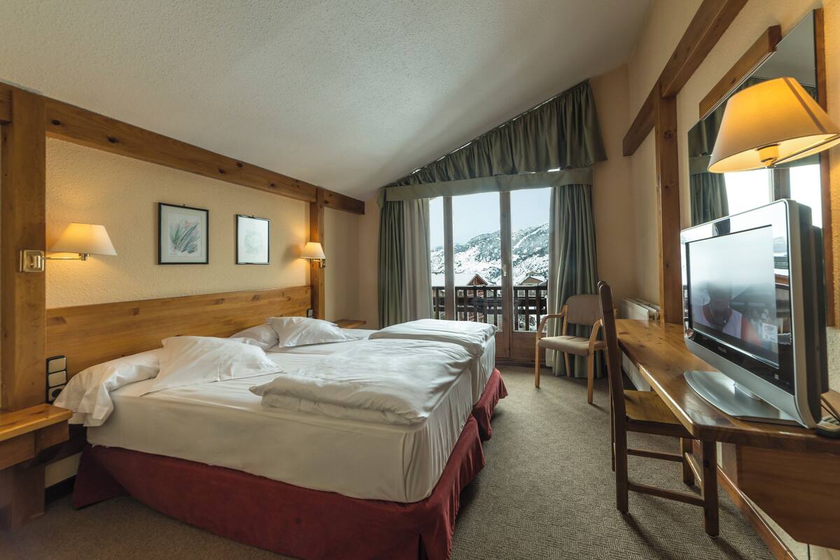 A hotel room with a wooden headboard, two beds with white linens, a television, and a view of snow-capped mountains through large windows.