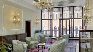 A spacious room with patterned armchairs, a red carpet, a chandelier, and a view of a courtyard through large windows. - Pausania Hotel (Venice)