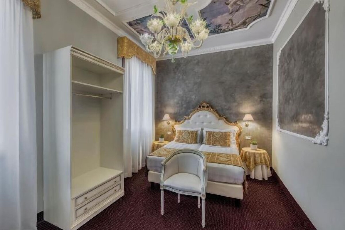 A hotel bedroom with a large bed, a wardrobe, a chair, and a chandelier.