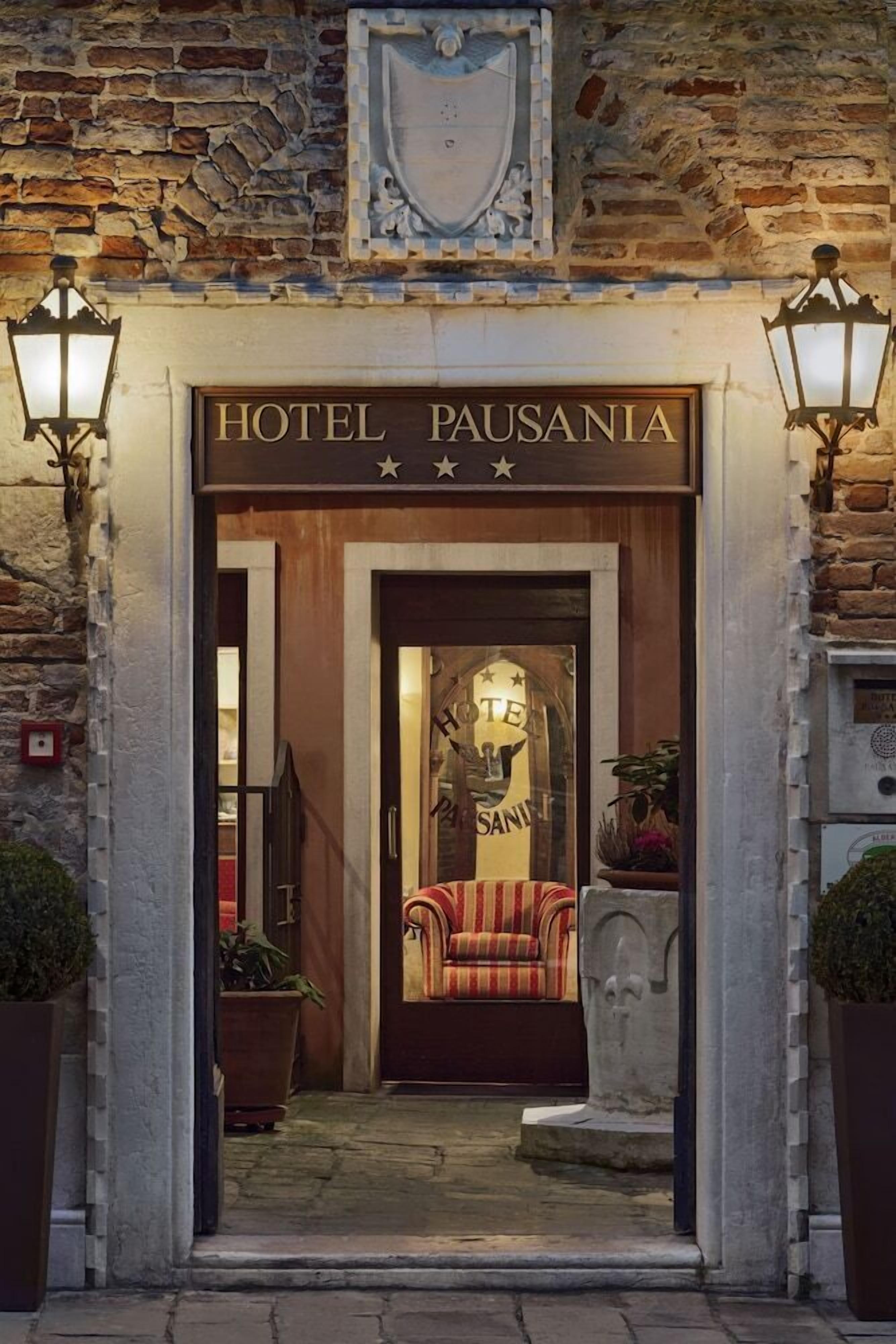 The entrance to Hotel Pausania with a wooden facade, a coat of arms above the entrance, and two wall-mounted lamps.