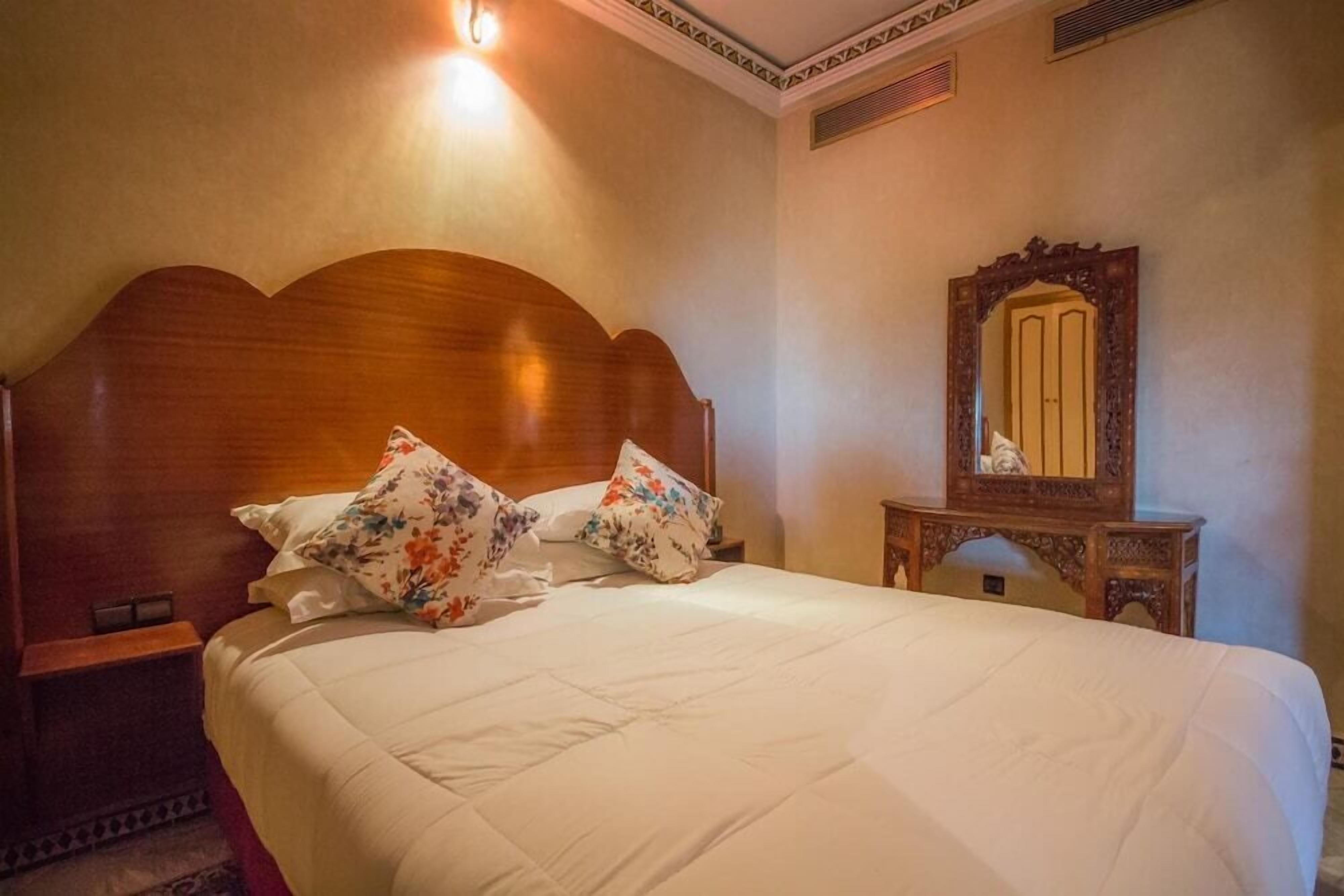 Una cama bien arreglada con almohadones florales, un cabecero de madera y una mesita de noche con espejo.