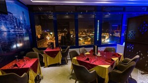 Un restaurante con mesas dispuestas para comer, amplias ventanas con vista a la ciudad y un mural de un paisaje urbano.