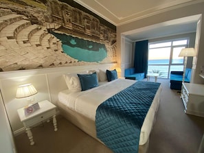 A hotel room with a large bed, a nightstand with a lamp, a wall mural, a sofa, and a view of the ocean. - Adriatik Hotel, BW Premier Collection (Durrës)