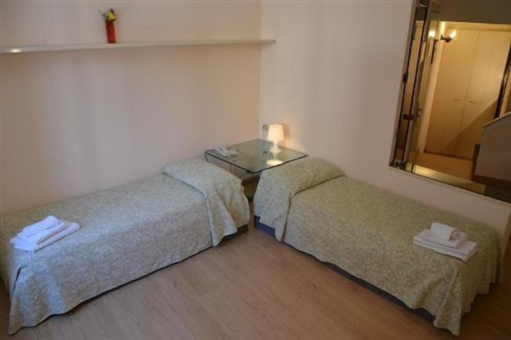 A small room with two single beds, a bedside table with a lamp, a mirror, and a shelf with a vase.
