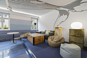 A room with a blue carpet, a round table, a sofa, a bean bag, a wooden chest, and a mural on the wall. - Hotel Britannia (Genoa)