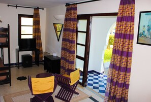 Ein Zimmer mit Schachbrettmuster-Boden, einem Fernseher, einem Schreibtisch, zwei Schaukelstühlen und violetten Vorhängen.