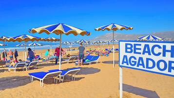 Una playa con múltiples sombrillas y sillas de descanso, un letrero que dice 'BEACH AGADOR', y un cielo azul despejado.