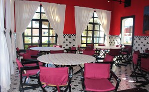 A dining area with round tables and red chairs, large windows with white curtains, and a red and black tiled floor. - Caribbean Village Agador (Agadir)
