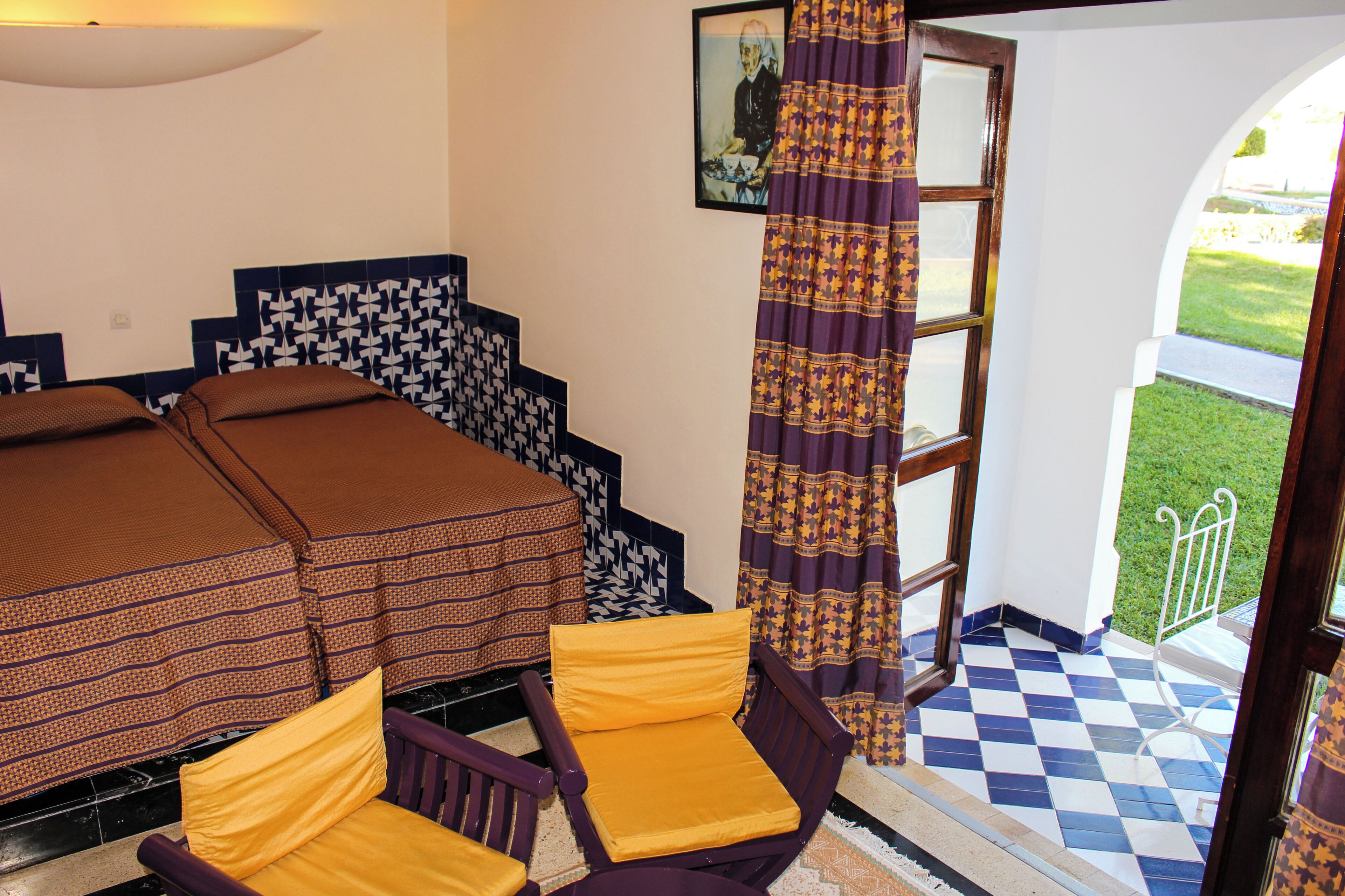 Une pièce comprenant un lit, deux chaises, un mur à motifs, une fenêtre avec des rideaux et un sol à carreaux.