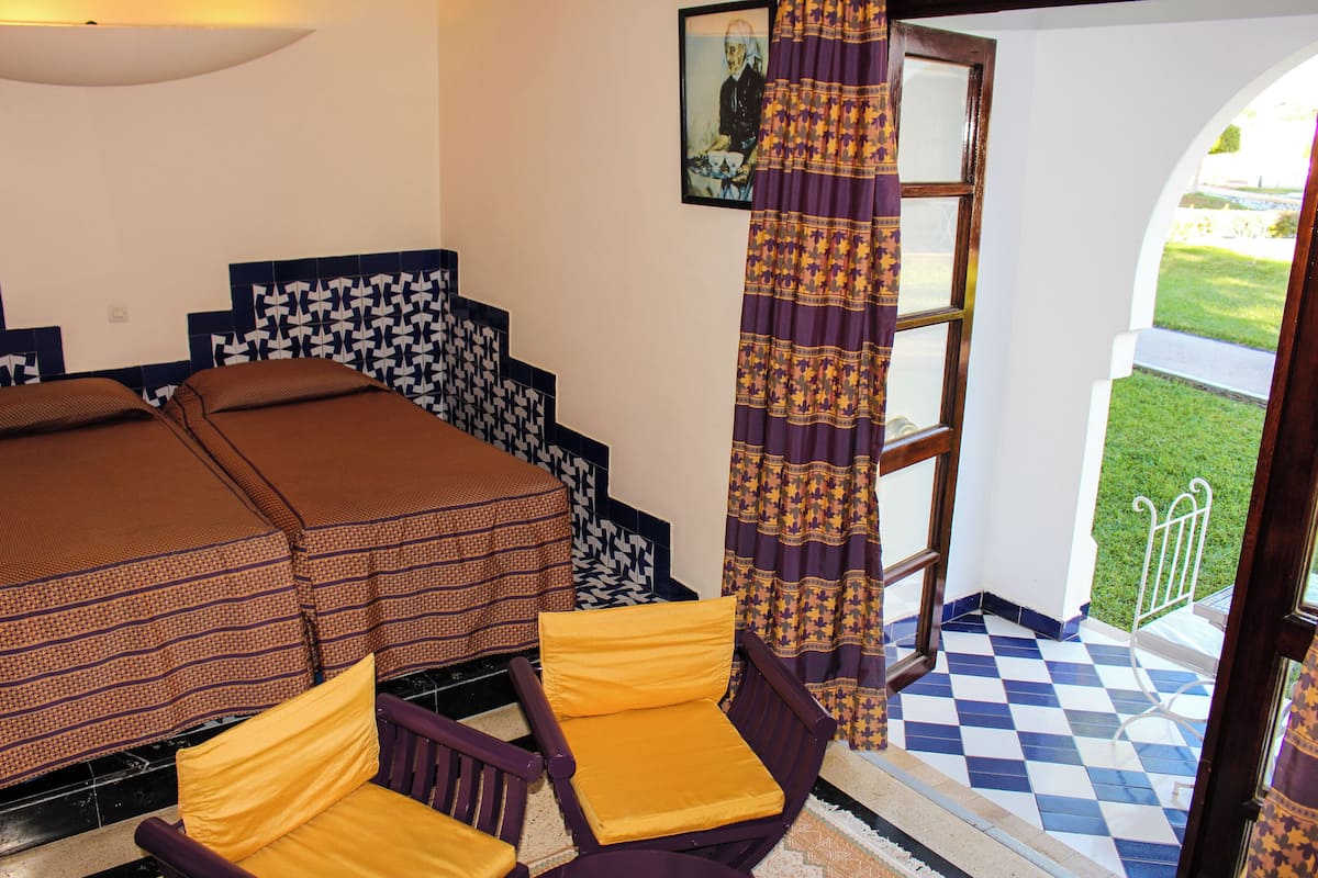 A room with a bed, two chairs, a patterned wall, a window with curtains, and a checkered floor.