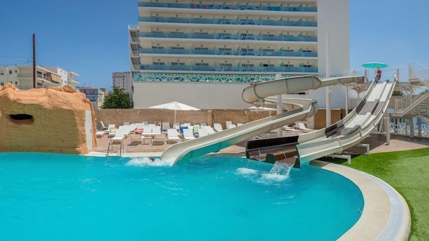 A pool with two water slides, lounge chairs, and a building in the background. - Villa Luz Family Gourmet & All Exclusive Hotel (Gandia)