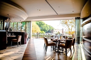 C'est un restaurant avec un long comptoir, plusieurs tables et des chaises. On peut voir des arbres et des bâtiments à travers les fenêtres.