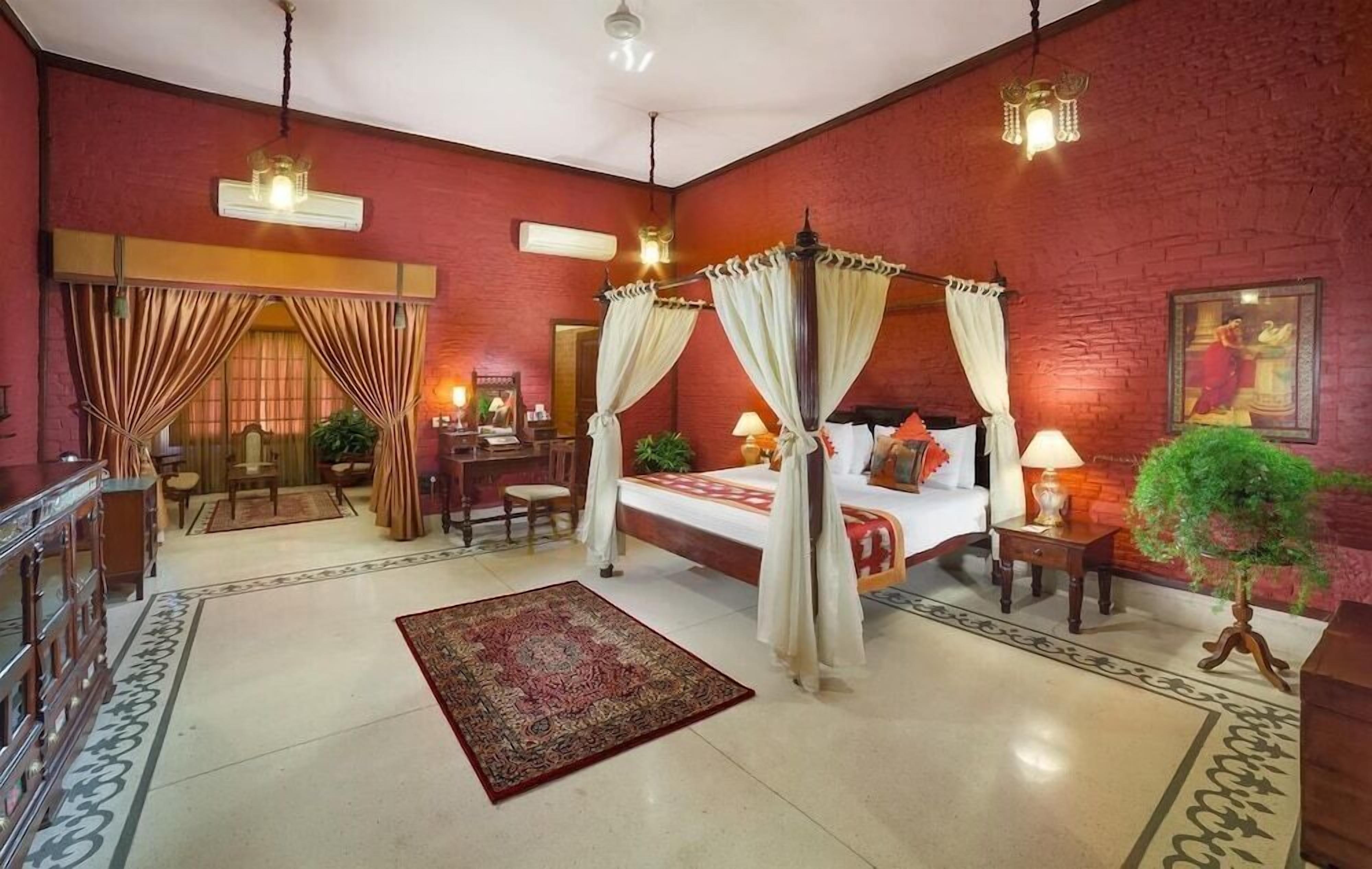 A spacious room with a large bed, a desk, and a chair. There are curtains, a rug, and a plant.