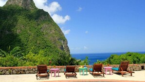 A terrace with wooden lounge chairs and small tables, overlooking a lush green mountain and the ocean. - Stonefield Estate Villa Resort (Soufrière)