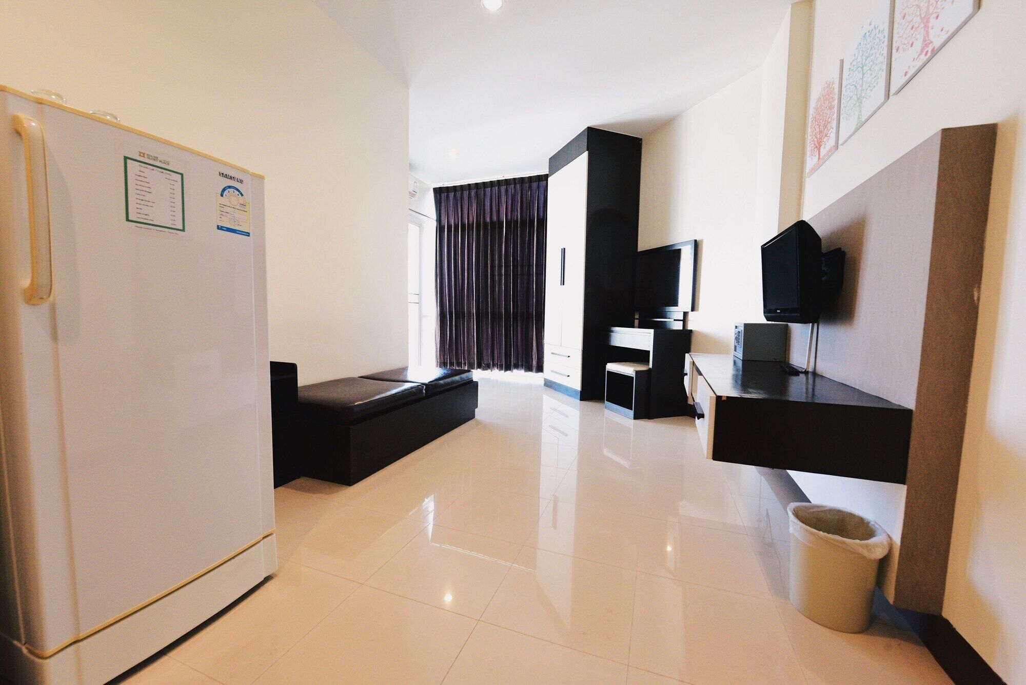 A modern hotel room with a refrigerator, a flat-screen TV, a desk, and a seating area with a bench.