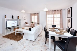 A modern living room with a white sofa, a dining table with black chairs, a TV on a white stand, and a large window with beige curtains. - The View Hotel Resort (La Oliva)