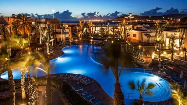 Piscina de un resort con palmeras y sillones de descanso, rodeada de edificios y un atardecer al fondo.