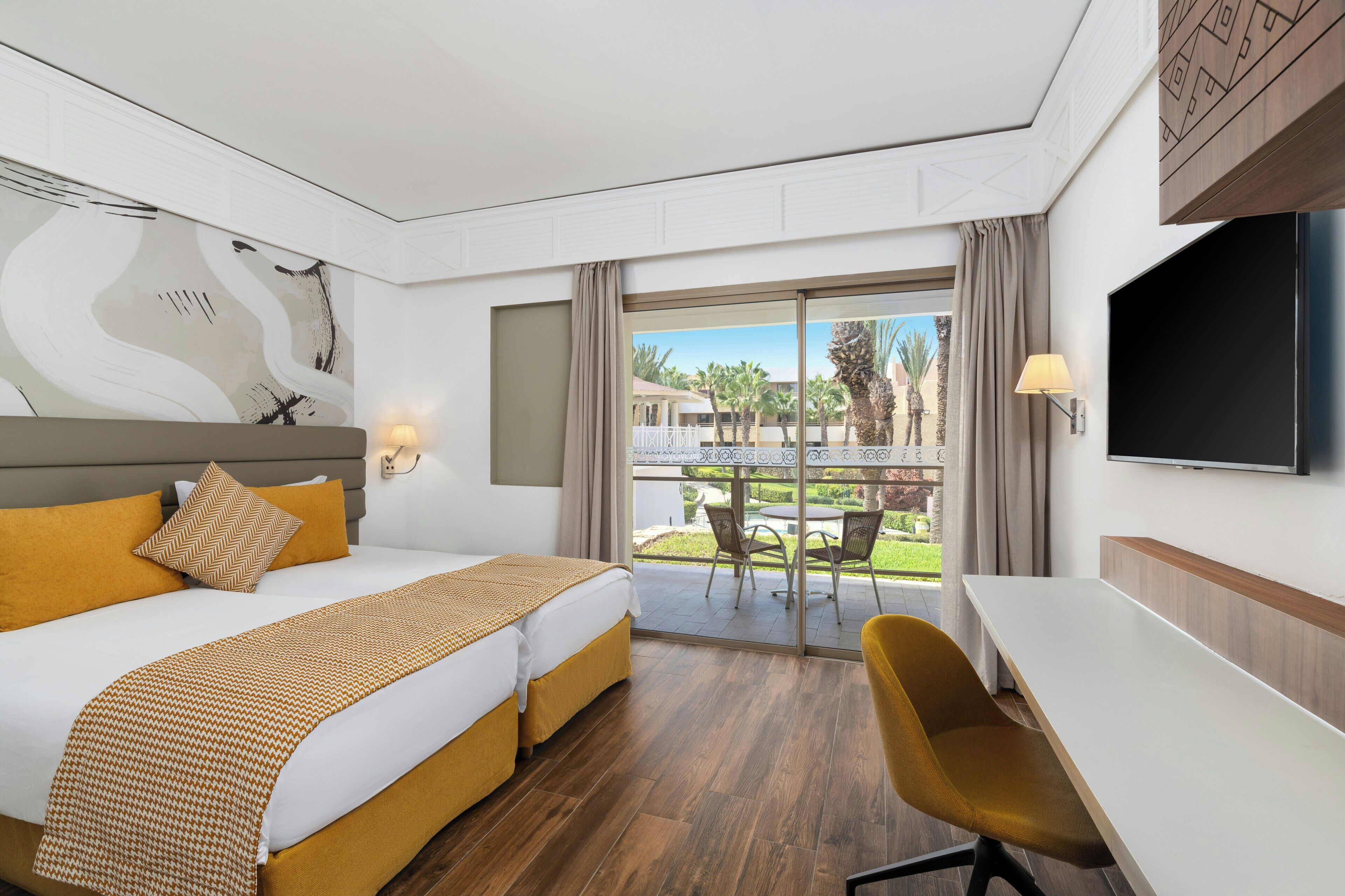 A hotel room with a bed, a desk, a chair, a TV, and a balcony with a view of palm trees.