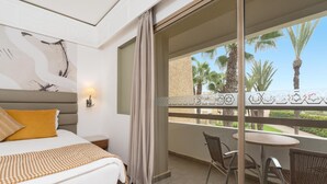 Habitación de hotel con cama, mesita de noche, lámpara, balcón con sillas de mimbre y una mesa, y vista a palmeras.