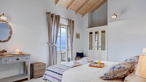 A bedroom with a bed, a nightstand, a dresser, and a view of the sea. - Villa Persephone (Kefalonia)