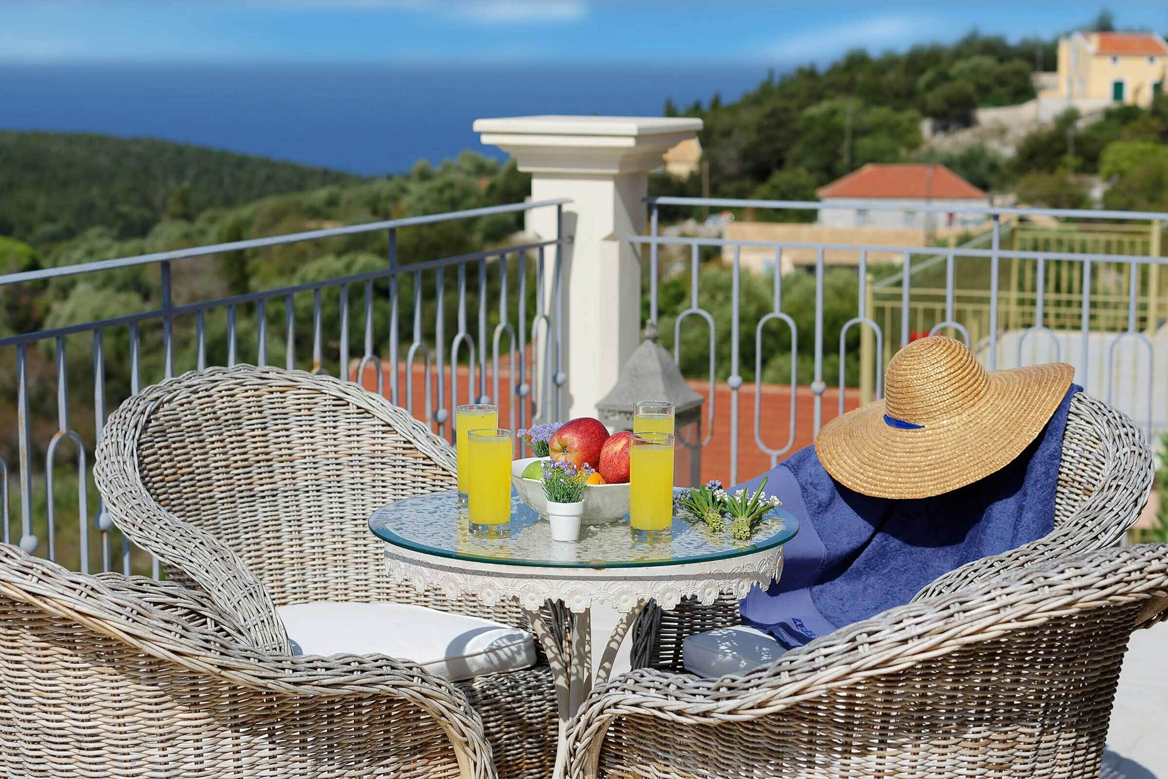 A wicker chair with a glass table, a white pedestal, and a view of the sea and hills.