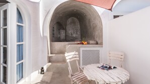 A white interior with a table and chairs, a small kitchen area, and a large arched doorway. - Alta Mare By Andronis (Santorini)