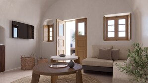 A cozy living room with a sofa, a round wooden table, a television mounted on the wall, and a large window. - Alta Mare By Andronis (Santorini)