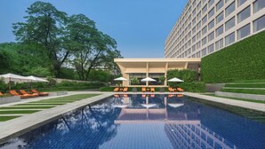 A swimming pool with lounge chairs and umbrellas, surrounded by greenery and a modern building.