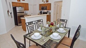 On aperçoit un coin repas avec une table en verre, dressée pour quatre personnes et entourée de chaises. Au fond, on distingue une cuisine avec des meubles en bois et un four à micro-ondes.