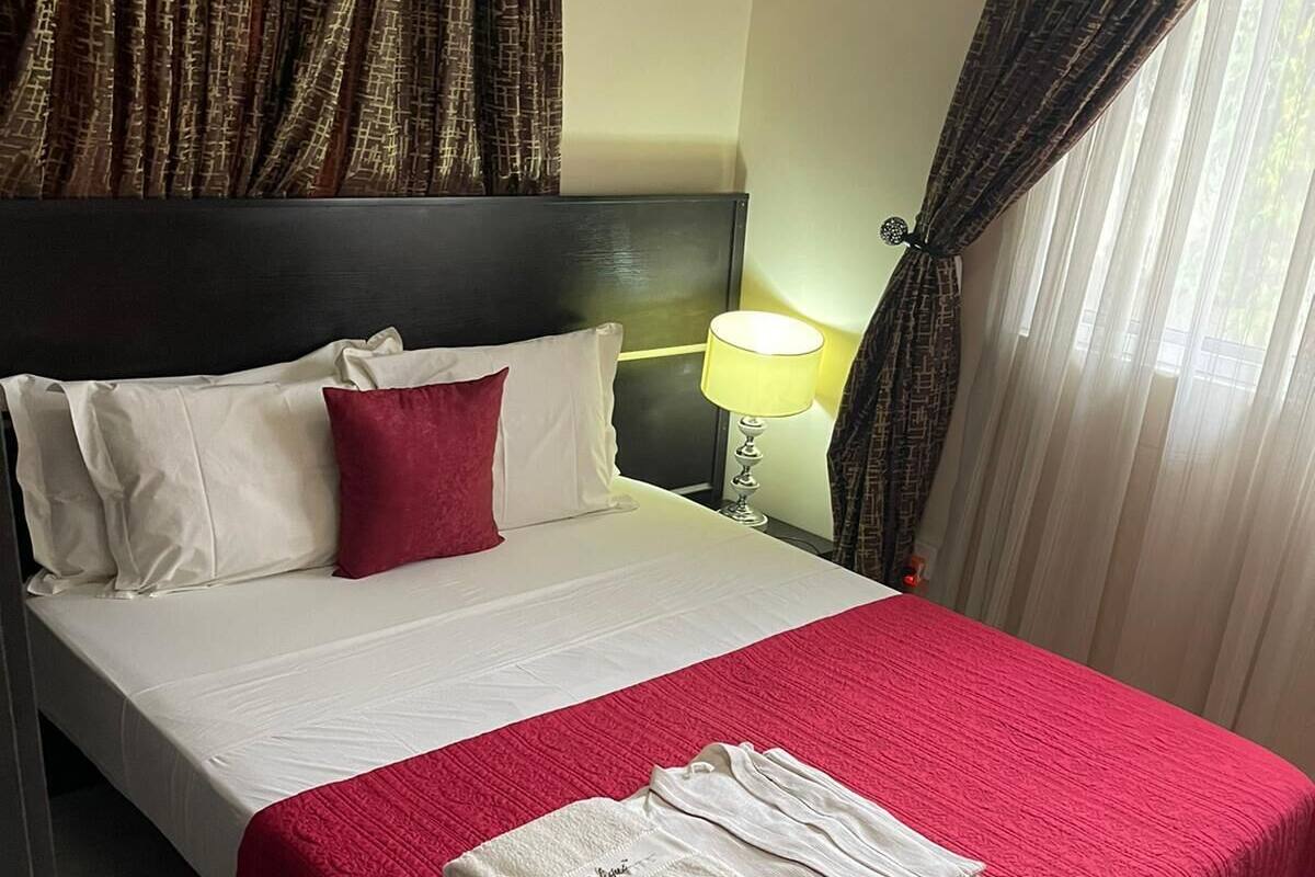 Una cama bien hecha con una colcha roja y ropa de cama blanca, una mesita de noche con una lámpara y una ventana con cortinas de gasa.