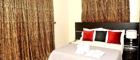 A hotel room with a bed, curtains, pillows, a bedside table with a lamp, and towels.