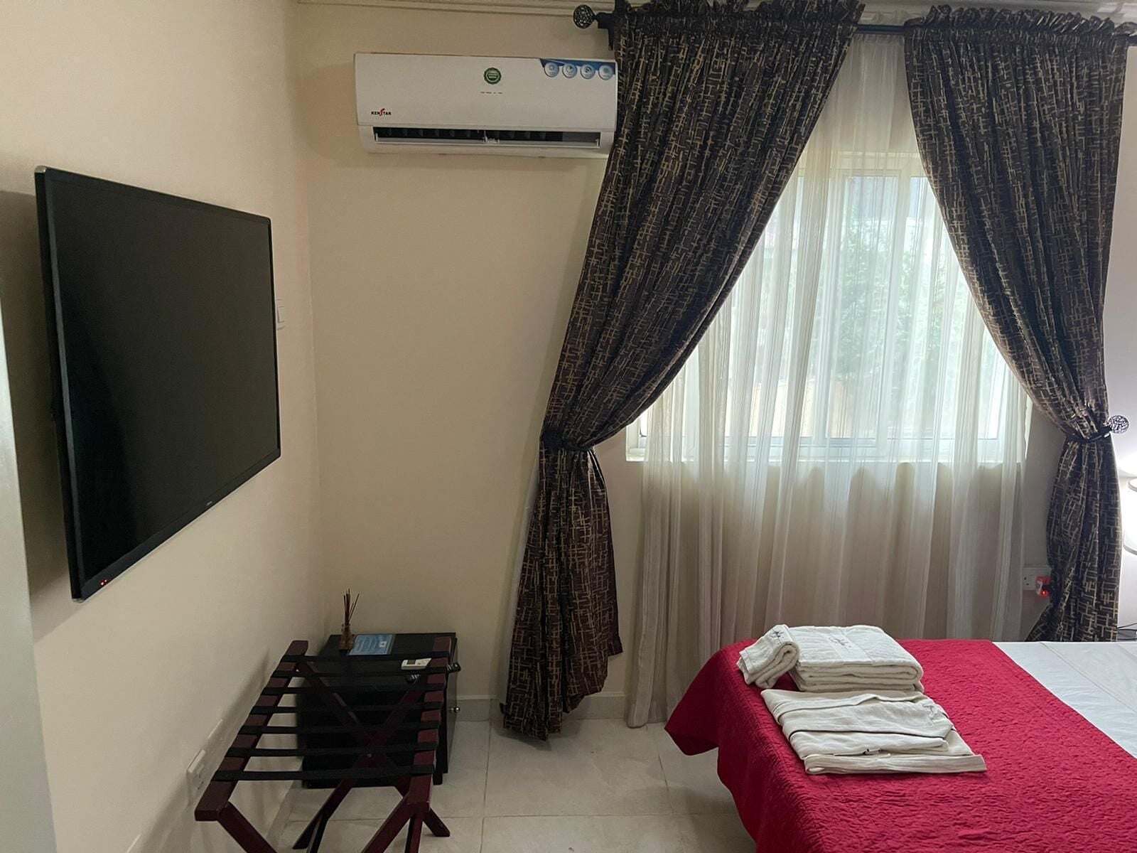Habitación de hotel con televisor de pantalla plana, aire acondicionado, una pequeña mesa con una silla, una cama con una manta roja y cortinas estampadas.