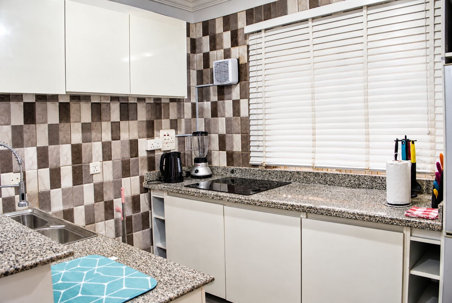 A kitchen with a checkered backsplash, a window with blinds, a sink, a stove, and a countertop with a kettle and a blender.
