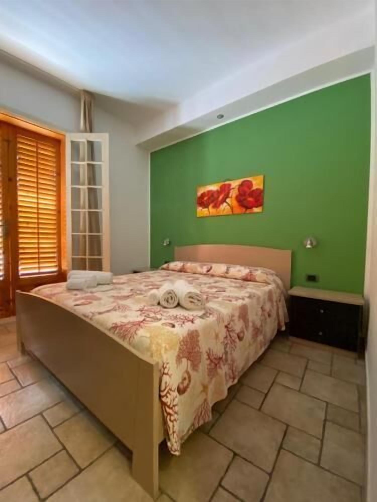 Een slaapkamer met een bed, een groene wand, een schilderij en een raam met jaloezieën.