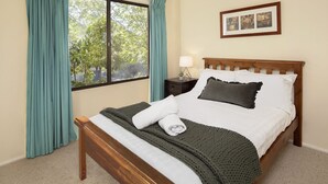 A neatly made bed with white linens, a dark grey throw blanket, and a white towel folded on top. There is a wooden nightstand with a lamp beside the bed. A framed picture hangs on the wall above the bed.