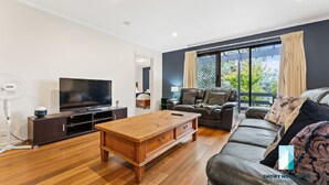 A modern living room with a flat-screen TV, wooden coffee table, leather sofa, and large windows with curtains.