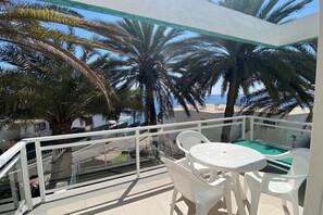 A balcony with a glass railing, white plastic chairs, a small round table, and a view of palm trees and the sea. - Acapulco (San Bartolomé de Tirajana)