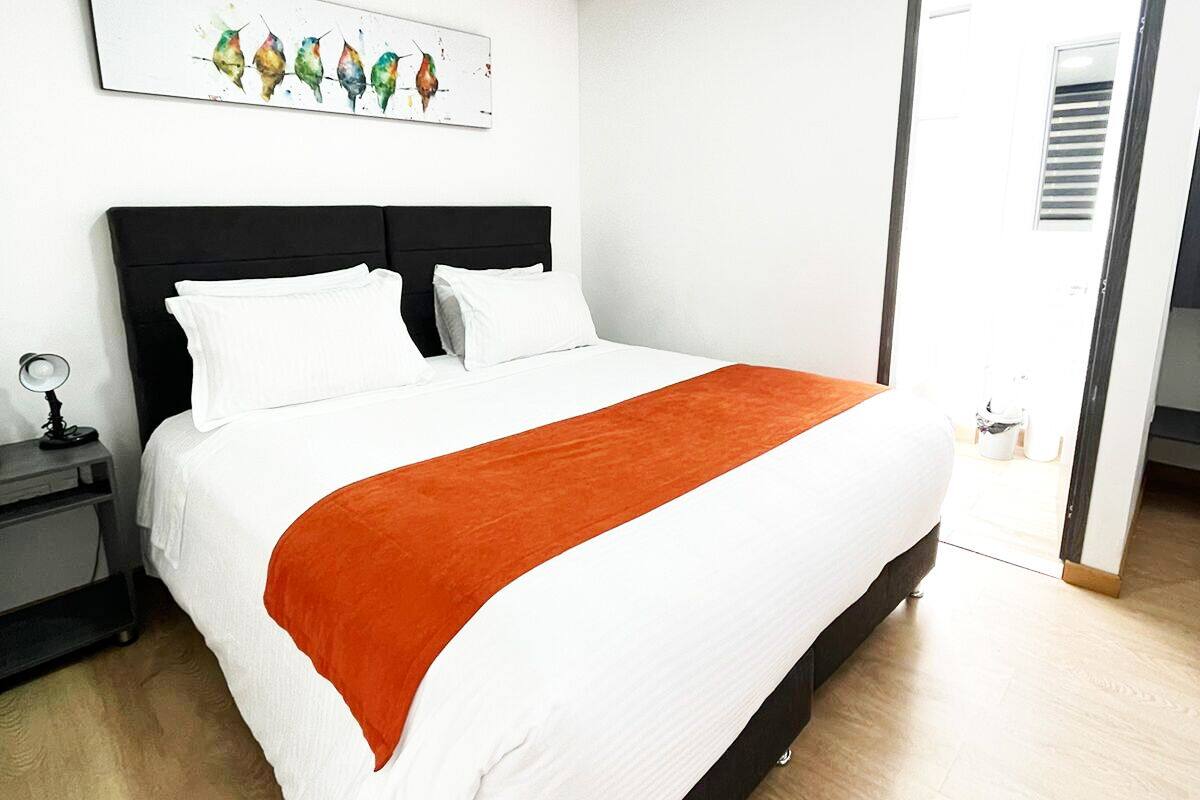 Un dormitori modern amb un llit gran, una tauleta de nit, una obra d'art penjada a la paret i una porta que dona a una altra habitació.