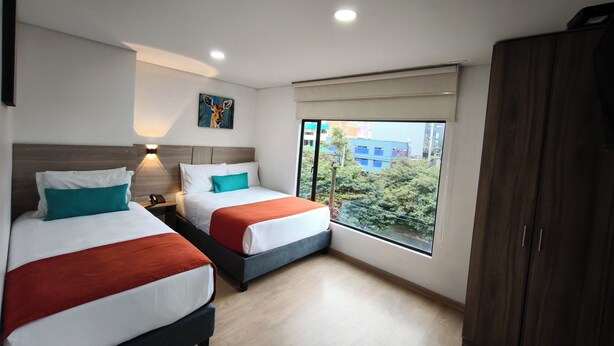 A hotel room with two beds, a nightstand, a painting, a window with a view of buildings, and a wardrobe. - Hotel Portal de los Andes (Bogotá)