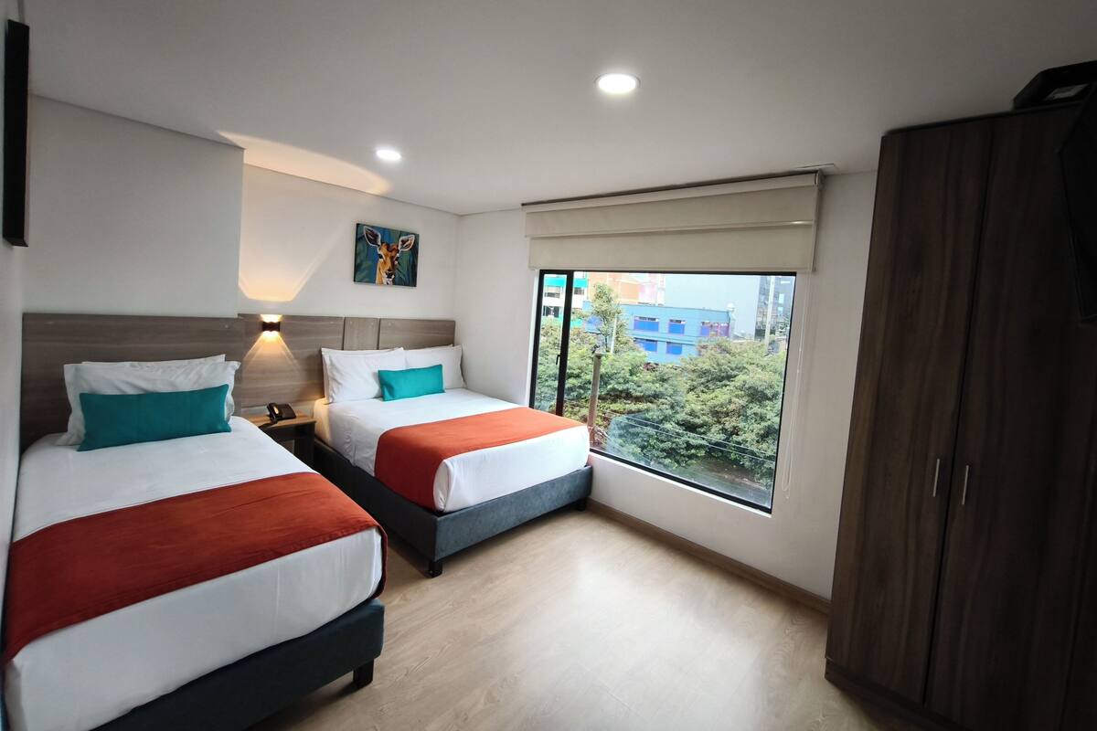 Habitació d'hotel amb dos llits, una taula de nit, un quadre, una finestra que mostra edificis i un armari.