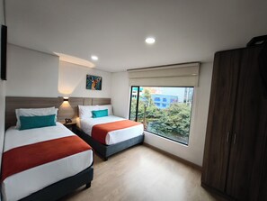 Ein Hotelzimmer mit zwei Betten, einem Nachttisch, einem Gemälde, einem Fenster mit Blick auf Gebäude und einem Kleiderschrank.
